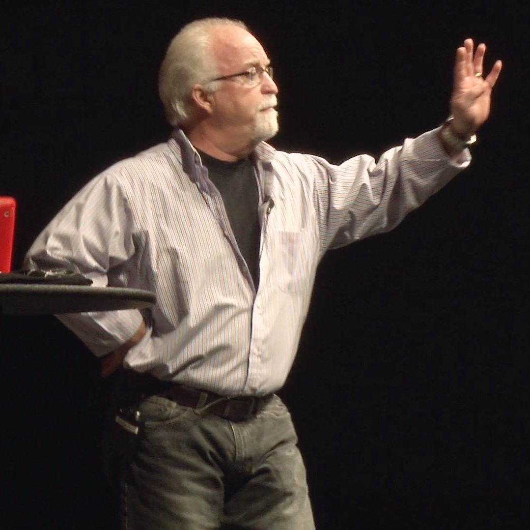 A man with white hair and glasses is holding his hand up.