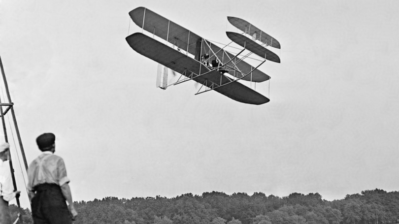 A black and white photo of an old biplane flying over trees.